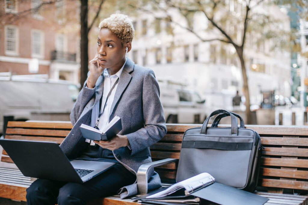 jeune femme avec des écouteurs dans les oreilles, elle est assise sur un banc avec son matériel : ordinateur, cahiers, notes